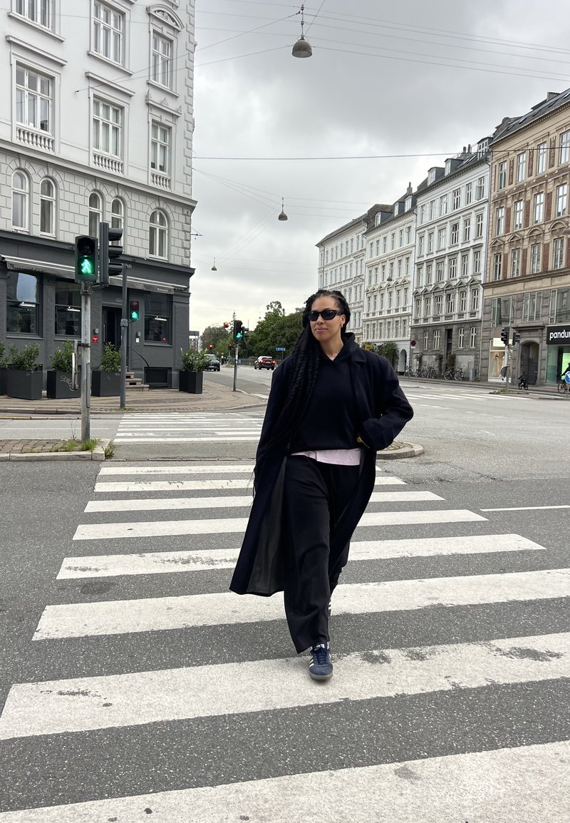 Cappotto lungo nero su una felpa nera e una maglietta rosa, abbinato a pantaloni neri e scarpe scure, in piedi su un attraversamento pedonale. Visibili strada e edifici.