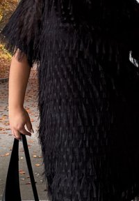 Černé texturované šaty s vrstvenými třásněmi, krátkými rukávy a volným střihem, doplněné černou kabelkou.