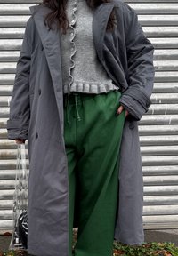 Casaco longo cinza, suéter cinza claro com bordas rendadas, calças verdes de corte descontraído, saco transparente com padrão preto e branco.