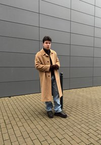 Μπεζ μάλλινο παλτό με μεγάλο γιακά, σκούφος από ζιβάγκο, μπλε τζιν και καφέ δερμάτινα παπούτσια με δέσιμο. Στέκεται κοντά σε μια μαύρη κολόνα σε πλακόστρωτο.