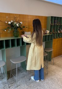 Geltonas vilnonis paltas ilgomis rankovėmis, su juosta ties liemeniu; žalia stiklinė vaza su gėlėmis ant žalio prekystalio; pilki taburetai ir tekstūruota grindų danga.