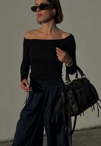 Femeie purtând un top negru cu umerii goi și ochelari de soare, ținând o geantă de piele neagră, stând în fața unui perete gri simplu, luminată de soare.