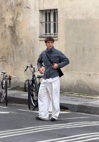 Hall hall mantel ees taskutega ja kõrge kraega, kombineeritud valgete laiade pükstega. Must õlakott ja tumedad kingad. Linnaline taust.