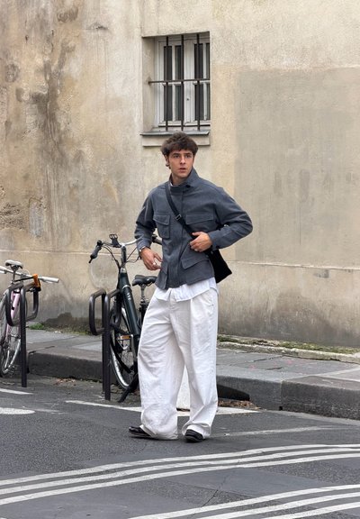 Chaqueta gris con bolsillos delanteros y cuello alto, combinada con pantalones blancos de pierna ancha. Bolso negro de hombro y zapatos oscuros. Fondo urbano.