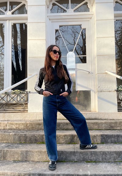 Chaqueta negra con cremallera y rayas blancas, combinada con pantalones vaqueros de mezclilla azul de tiro alto. Zapatillas negras con detalles blancos. Gafas de sol y un bolso de estampado de leopardo.