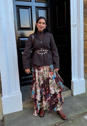 Chaqueta marrón de mezcla de lana sobre un cuello alto marrón ajustado, combinada con una falda floral en capas en tonos granate y crema, y botas marrones con tacón.