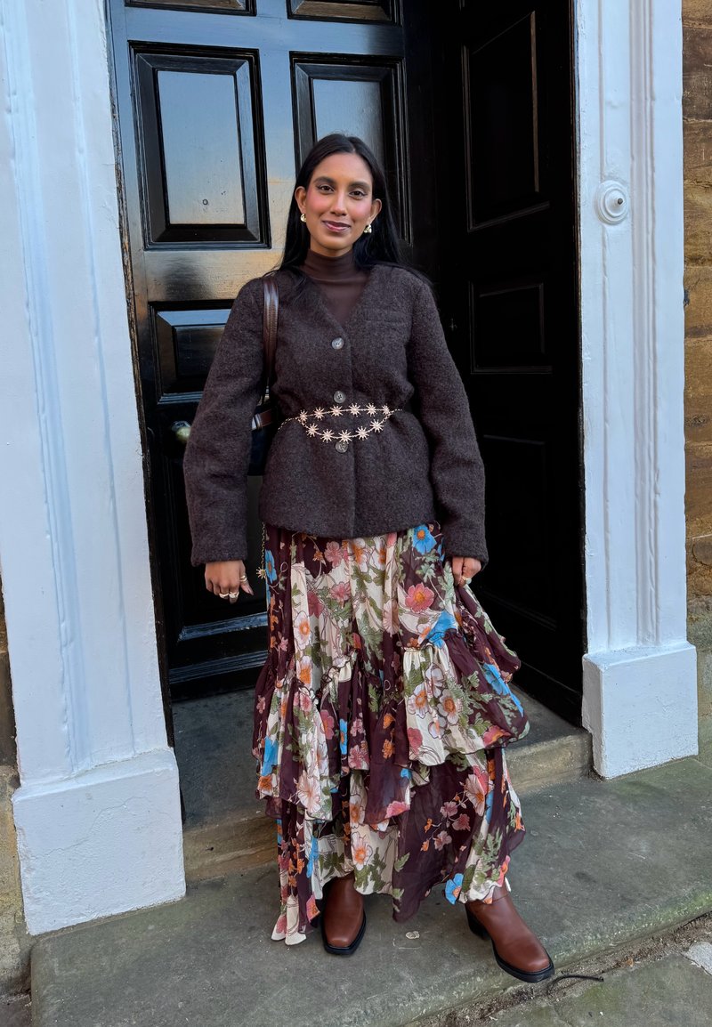 Veste en mélange de laine marron portée sur un col roulé marron ajusté, associée à une jupe superposée à fleurs en bordeaux et crème, et des bottes à talons marron.