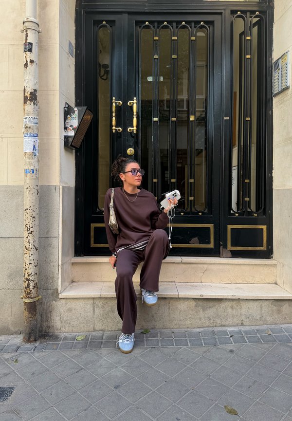 Mujer con ropa marrón oscuro y zapatillas deportivas sentada en un saliente de piedra frente a puertas dobles de metal negro, sosteniendo un teléfono inteligente y auriculares.
