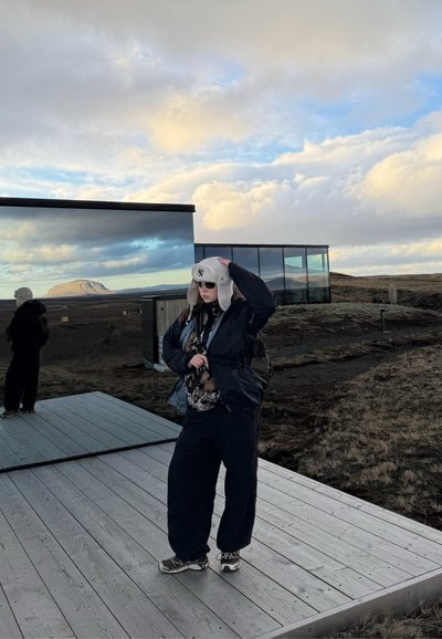 Persona con ropa de invierno y gorro de pie sobre una plataforma de madera junto a un edificio de vidrio reflectante en un paisaje abierto y nublado con colinas al fondo.