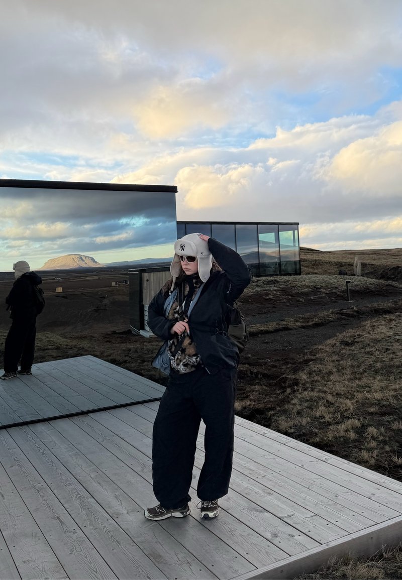 Person in Winterkleidung und Mütze steht auf einer Holzplattform neben einem reflektierenden Glasgebäude in einer offenen, wolkigen Landschaft mit Hügeln im Hintergrund.