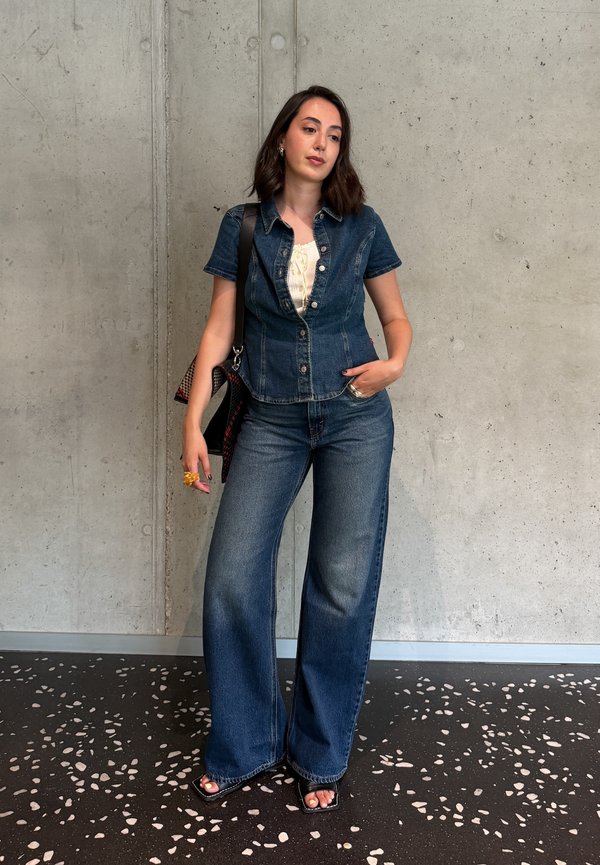 Femme debout contre un mur en béton, portant une chemise en jean à manches courtes boutonnée, un jean à jambes larges, des sandales noires et portant un sac à bandoulière.