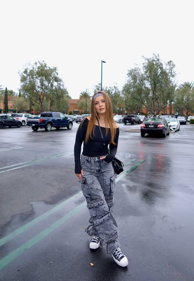 Top de manga larga negro combinado con pantalones cargo grises que cuentan con múltiples bolsillos y correas. Zapatillas blancas completan el atuendo. Al aire libre, pavimento mojado.