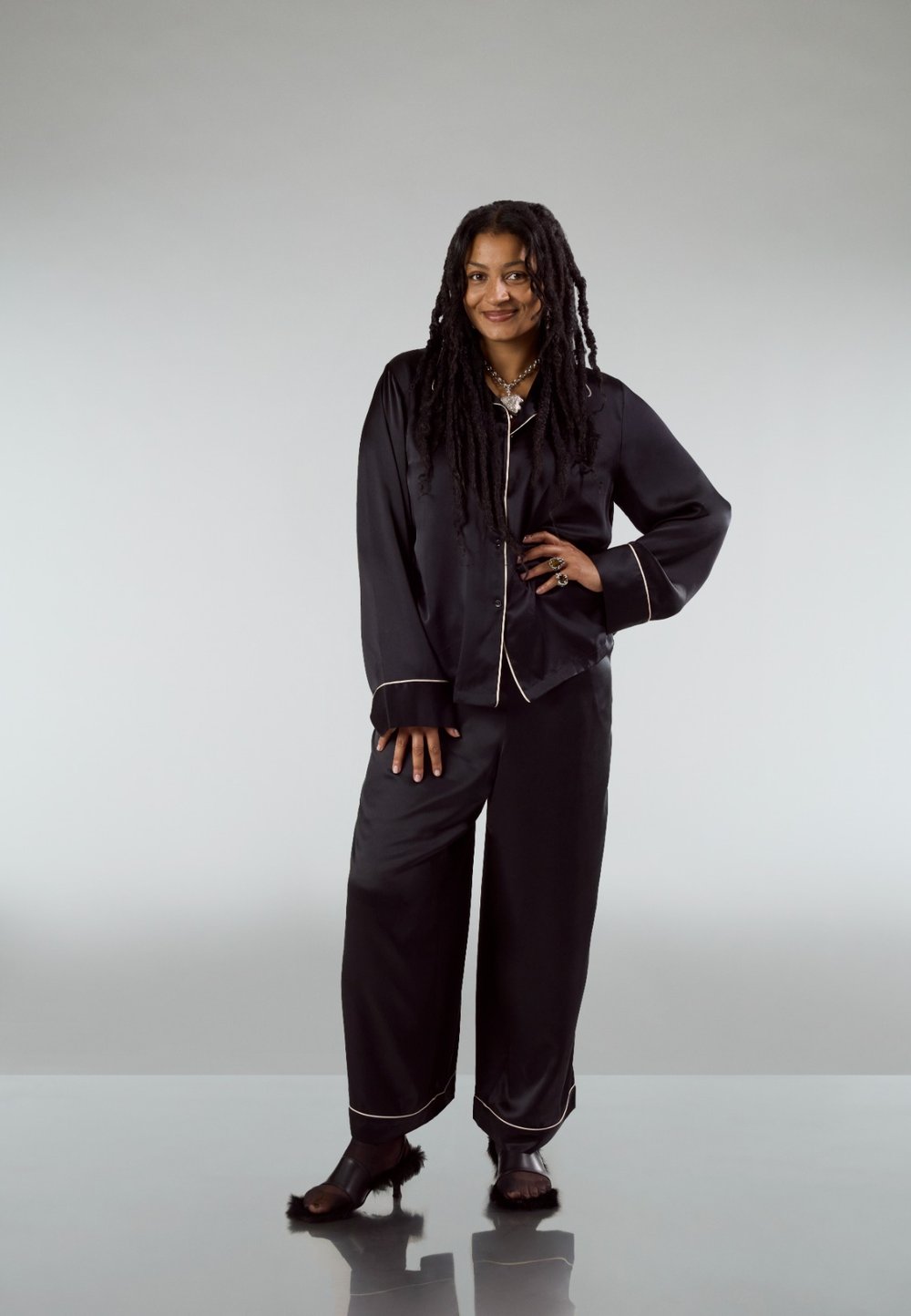 Zwart satijnen pyjamaset met witte biezen, bestaande uit een overhemd met knopen en broek met wijde pijpen, aangevuld met chunky zwarte sandalen.