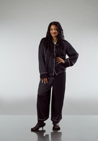 Zwart satijnen pyjamaset met witte biezen, bestaande uit een overhemd met knopen en broek met wijde pijpen, aangevuld met chunky zwarte sandalen.