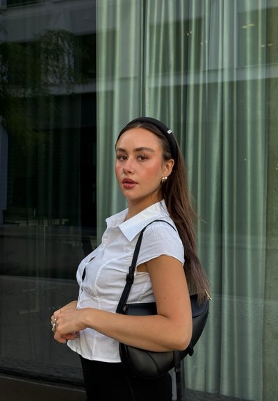 Bolso negro de hombro llevado cruzado, camisa blanca de manga corta con botones, pelo largo, con una diadema negra, frente a un fondo de cortina verde.