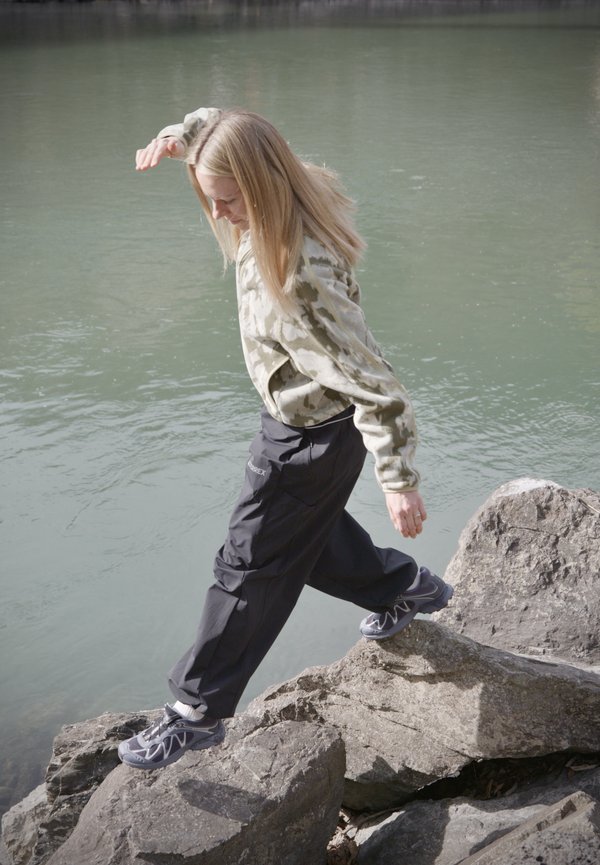 Junge Person mit blondem Haar, die vorsichtig auf großen Felsen nahe ruhigem Wasser geht und eine Tarn-Sweatshirt sowie dunkle Hosen trägt.