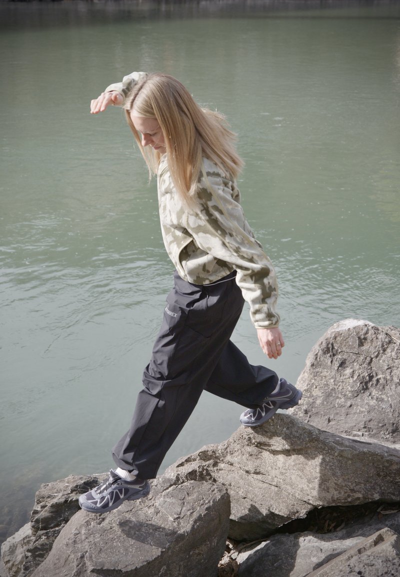 Jeune personne aux cheveux blonds marchant prudemment sur de gros rochers près d'une eau calme, portant un sweat-shirt camouflage et un pantalon sombre.