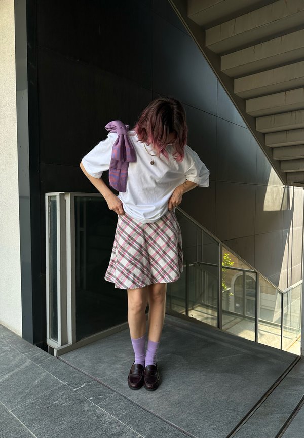 T-shirt bianco abbinato a una gonna a campana a quadri rosa. Calzini viola e mocassini scuri completano il look. Capelli leggermente ondulati con riflessi rosa.