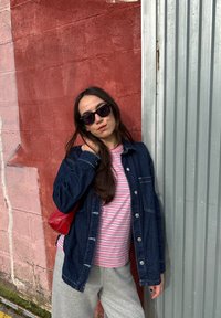 Chaqueta vaquera, camiseta de rayas rosas y blancas, pantalones de chándal grises, gafas de sol y bolso rojo. Fondo urbano con paredes texturizadas.