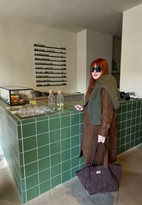 Casaco de camurça castanho emparelhado com um suéter verde, segurando uma bolsa tote escura. Balcão de azulejos verdes com garrafas de água de vidro e pastelaria em exibição.