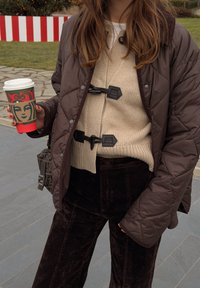 Brązowa pikowana kurtka z czarnymi elementami, kremowy sweter prążkowany i ciemnobrązowe spodnie sztruksowe, trzymająca świąteczny kubek kawy.