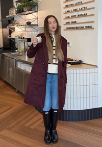 Kvinne med langt hår iført maroon-frakk og svarte gummistøvler holder en kopp, poserer i en kafé med en meny tavle og bakverk bak seg.