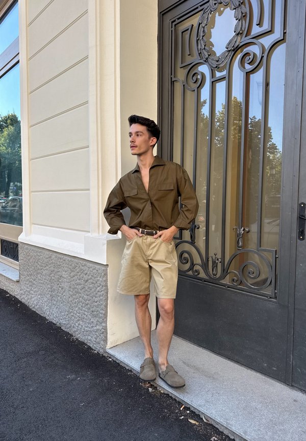 Un hombre con una camisa marrón y pantalones cortos beige está de pie frente a una puerta negra ornamentada y mira hacia un lado.