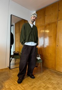 Homme en cardigan vert, chemise blanche, bonnet beige et pantalon marron debout près d'une chaise verte sur laquelle un chat est assis, à l'intérieur d'une pièce avec des armoires en bois.