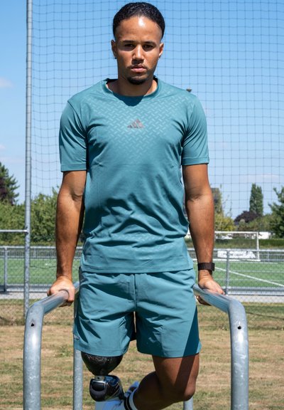 Hombre con pierna prostética vestido con atuendo deportivo en color azul verdoso haciendo fondos en barras paralelas en un campo de deportes al aire libre bajo un cielo despejado.