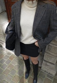 Szürke kockás blézer krémszínű garbóval, fekete miniszoknyával és térdig érő fekete csizmával párosítva. Fekete válltáska. Kőköves háttér.