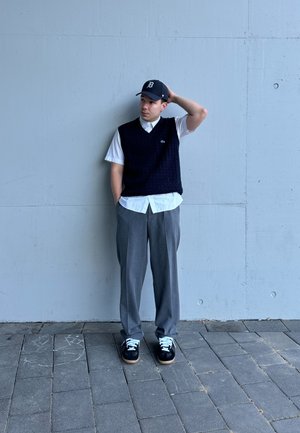 Chaleco de punto azul marino sobre una camisa blanca con cuello, combinado con pantalones grises y zapatillas negras con cordones blancos, de pie frente a una pared de concreto.