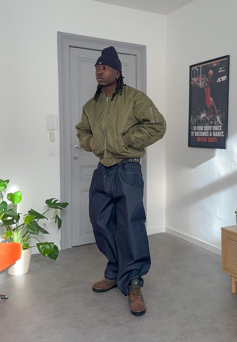 Homme portant une veste bomber verte, un bonnet bleu marine, un jean ample et des bottes marron, debout dans une pièce avec des plantes et une affiche de motivation de basketball.