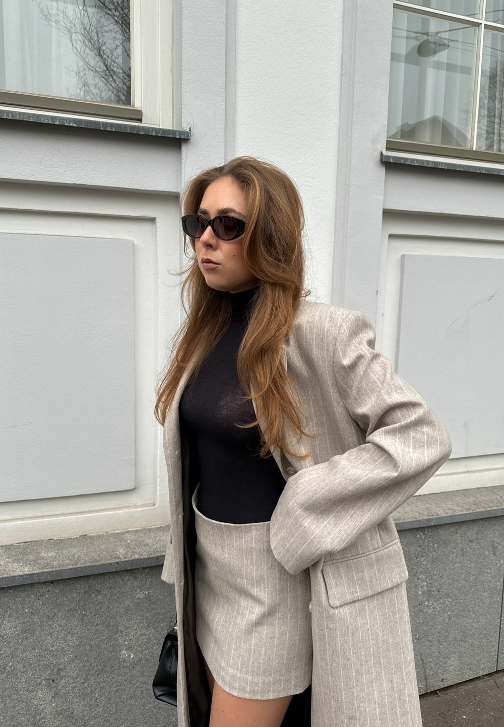 Blazer largo de rayas en gris claro sobre un suéter negro de cuello alto, combinado con una minifalda a juego. Los accesorios incluyen gafas de sol oscuras y un pequeño bolso negro.