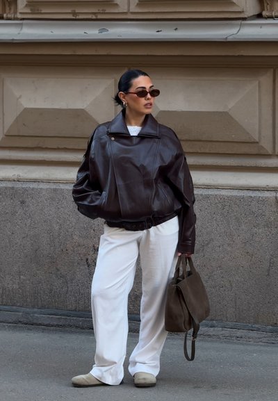 Mujer con chaqueta de cuero marrón oscuro, pantalones blancos, zapatos beige, sosteniendo un bolso marrón, de pie frente a la fachada de un edificio de piedra.