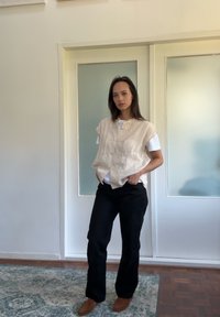 Tunică bej texturată, cu mâneci scurte, suprapusă peste un tricou alb, asociată cu pantaloni negri și pantofi maronii, pe un fond deschis.