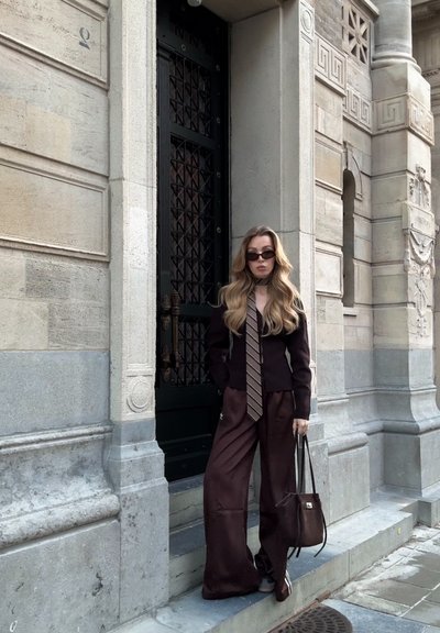 Blazer ajustado marrón oscuro con corbata a rayas en contraste, pantalones de piernas anchas y bolso a juego. Tela suave, diseño elegante y gafas de sol.