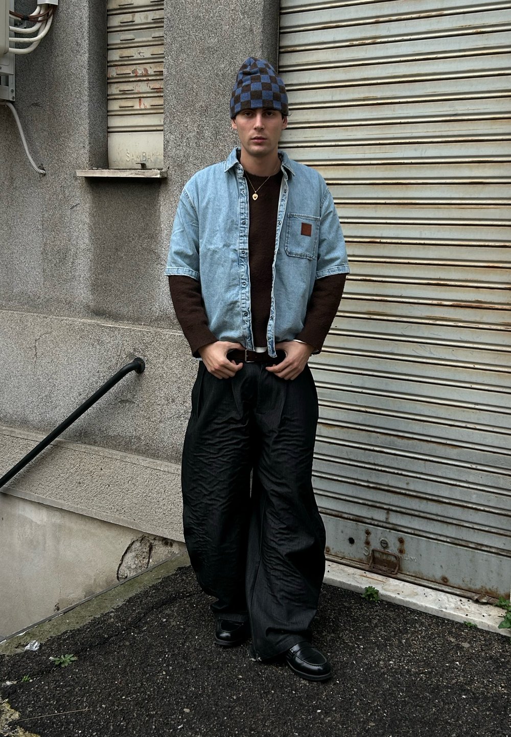 Chemise en jean bleu clair portée sur un pull marron, associée à un pantalon large texturé noir et un bonnet à carreaux bleu et noir. Chaussures noires.