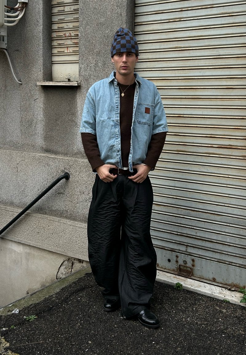 Lyseblå denimskjorte over en brun sweater, kombineret med sorte, strukturerede bukser med vide ben og en blå- og sortternet hue. Sorte sko.