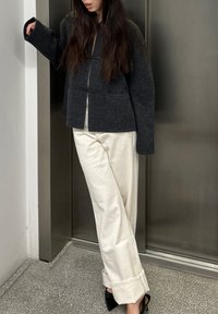 Veste en laine gris foncé à coupe carrée, fermée par un bouton à bascule, associée à un pantalon large crème et des talons noirs, sur un fond métallique.