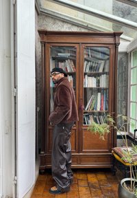 Chaqueta de piel de peluche marrón, pantalones de cuero oscuros y mocasines con estampado, de pie junto a una estantería de madera con puertas de cristal en una habitación luminosa.