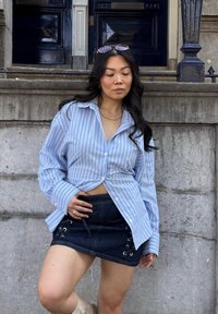 Helleblaue gestreifte Hemdbluse mit lockerem Schnitt, kombiniert mit einem dunklen Jeans-Minirock, der Schnürdetails und einen seitlichen Kordelzug hat.