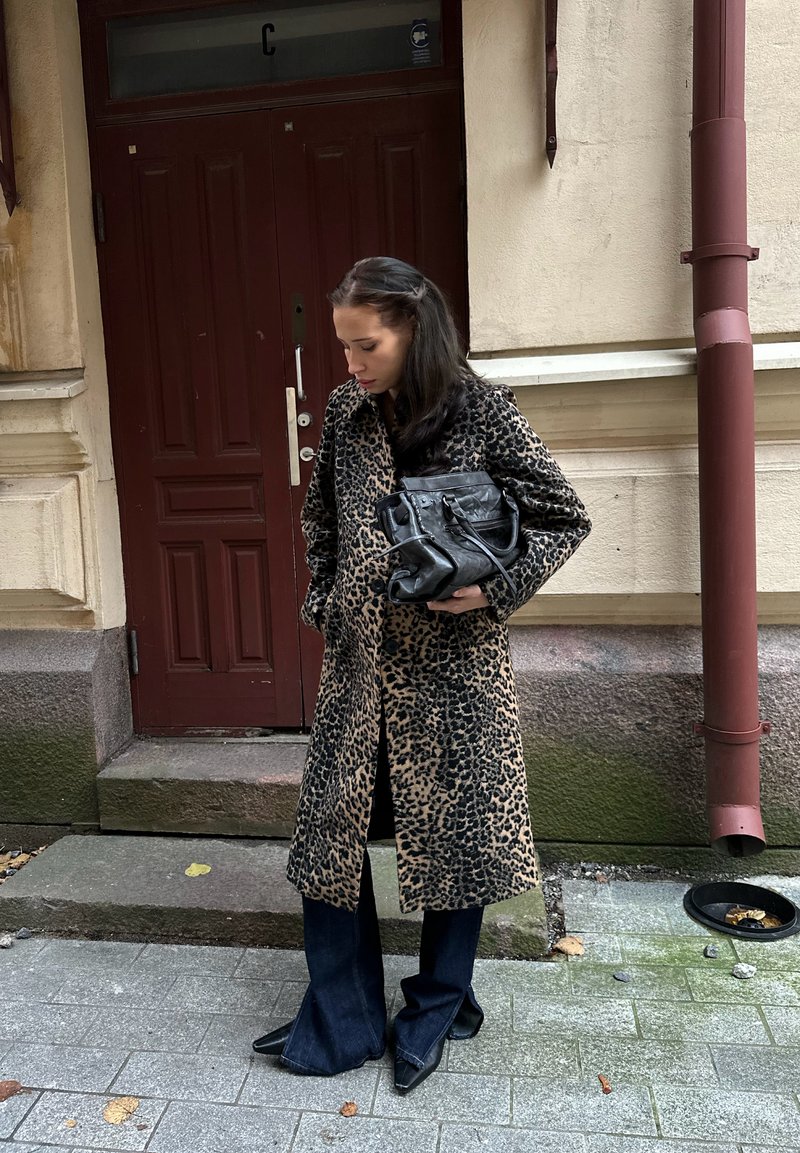 Casaco trench coat com estampa de leopardo, mangas compridas, parte da frente aberta. Calças de ganga escuras flare. Saltos pretos pontiagudos. Mala preta com alça. Fundo de pavimento de pedra.