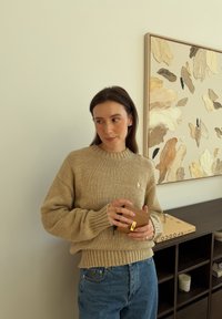 Γυναίκα με μπεζ πουλόβερ και μπλε τζιν κρατά ένα μικρό καφέ πορτοφόλι, στέκεται κοντά σε ένα σκοτεινό ράφι με διακοσμητικά και αφηρημένη τοιχογραφία.