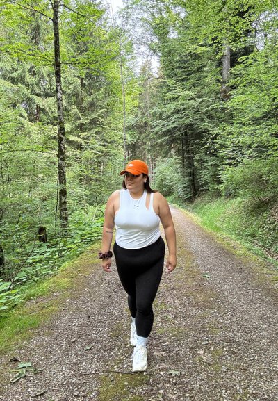 Persona caminando por un sendero de grava en un bosque, vistiendo una camiseta sin mangas blanca, leggins negros, una gorra naranja y zapatillas deportivas blancas con calcetines estampados.