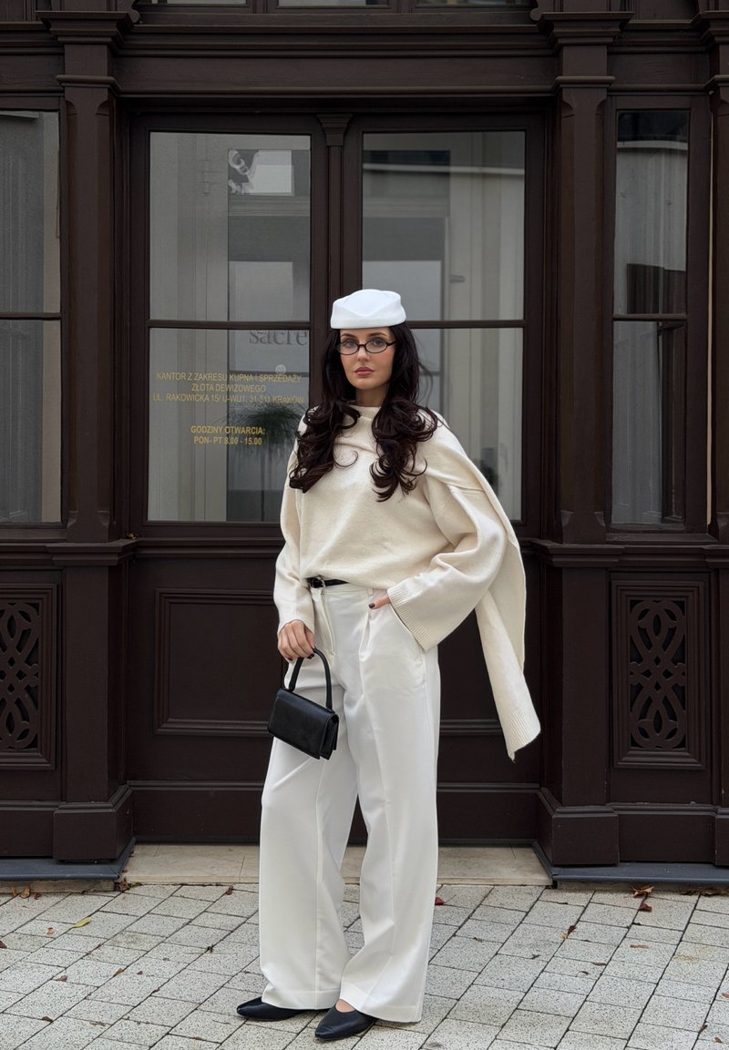Bela pletena jopica, široke bele hlače, črna torbica, črne balerinke in bela kapa s očali. Gladke teksture, nevtralna barvna paleta.