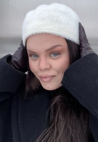 Jeune femme aux yeux bleus portant un bonnet blanc en tricot, un manteau noir et des gants en cuir foncé, tenant son bonnet par une journée froide.