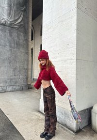 Pull court texturé rouge à manches larges, associé à un pantalon camouflage. Le mannequin tient un parapluie, debout contre un mur en pierre.
