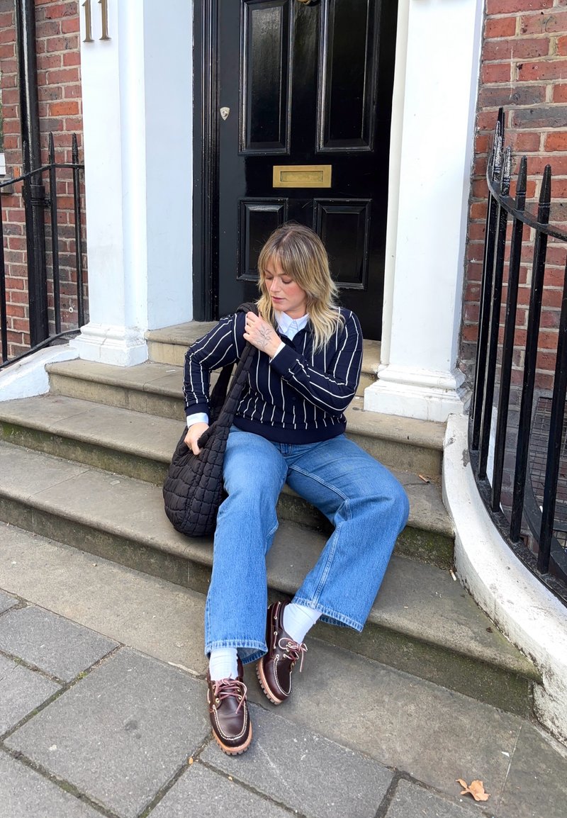 Pullover a righe blu navy, jeans a gamba larga blu, scarpe marroni scure e una borsa trapuntata nera. Ambientazione: gradini di pietra accanto a una porta nera con un numero.