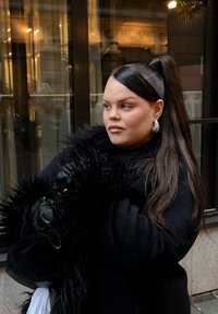 Sort pelsfrakke med lange ærmer, kombineret med sølvfarvede hoop-øreringe. Langt, glat hår og holder sorte solbriller. Baggrunden indeholder glas.