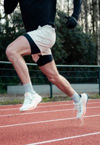Valkoiset urheilushortsit mustilla yksityiskohdilla, harmaat urheilusukat ja valkoiset juoksukengät oransseilla yksityiskohdilla. Aihe on puolivälin askeleessa radalla.